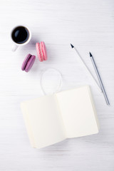 A cup of coffee, macarons, notebook and pencil on the white wooden table.