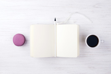 A cup of coffee, macaron, notebook and pencil on the white wooden table.