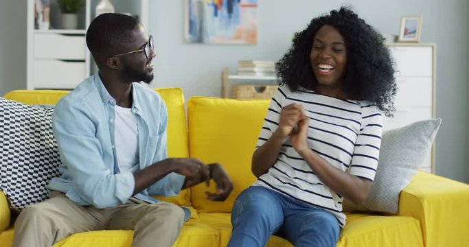 Portrait shot of the happy laughing African American couple tickling each other on the yellow couch and laughing laud. Indoors.