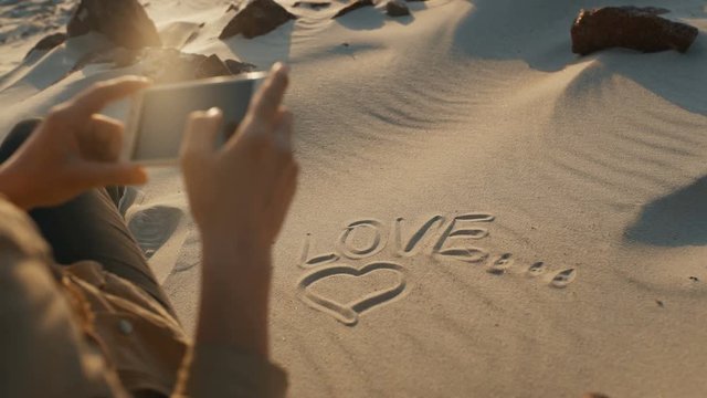 A girl takes a picture on the phone of an inscription on the sand "I love". Girl draws a symbol of the heart and love on the sandy beach, rear view, close-up. The concept of love. 4k