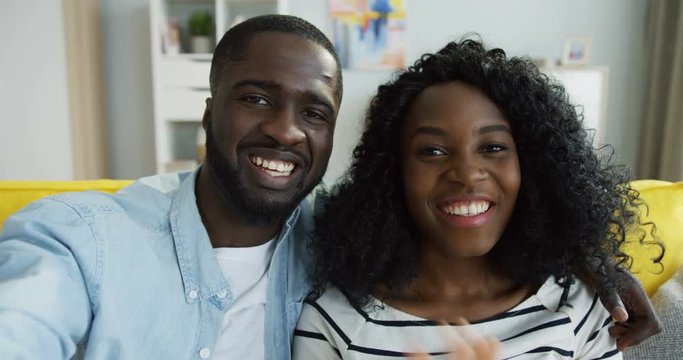 Close up of the young attractive African American man and woman videochatting joyfully and waving hands while sitting on the yellow couch at home. POV. Inside.
