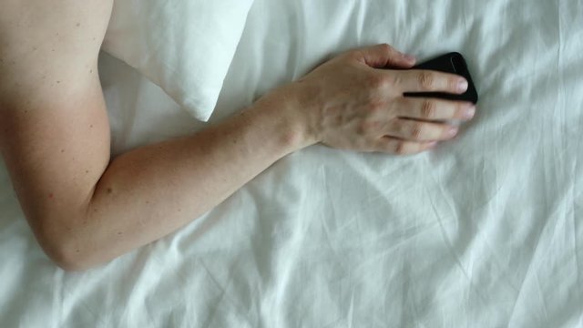 TOP VIEW: Human hand takes a phone on a bed and disable it in the morning