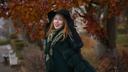 Red-haired girl in autumn park. She's wearing short coat and wide-brimmed hat. Woman is walking.