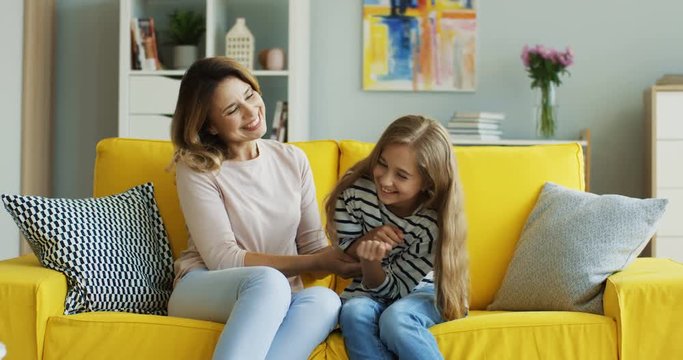 Cheerful Caucasian woman and her cute teen daughter sitting on the yellow couch, laughing and tickling. At home. Inside.
