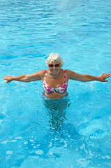 Fototapeta na wymiar Aged woman is doing spa exercises in bright blue water of pool.