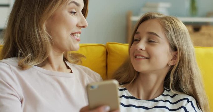 Close up of the Caucasian smiled mother and daughter resting on the sofa at home and watching something on the smartphone and talking. Indoor.
