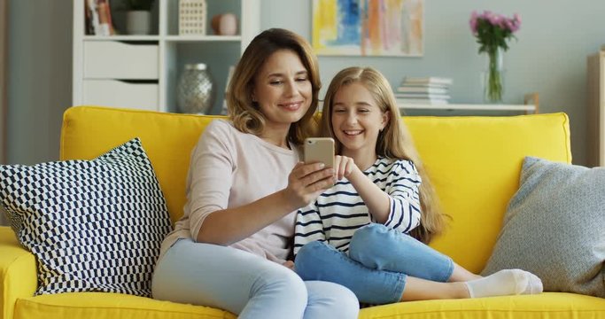 Beautiful blonde mother and pretty teenage girl sitting on the yellow sofa and watching something on the smartphone. Inside.