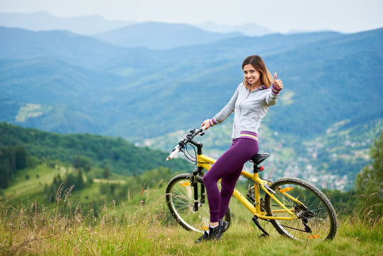 Smiling sporty female biker standing with yellow mountain bike on the top of a mountain, showing thumbs up sign. Mountains, forests and small city on the blurred background. Outdoor sport activity