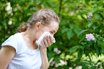 Fototapeta premium Allergy. Woman blowing her nose when standing close to flowers in a park. Woman with infection sneezing