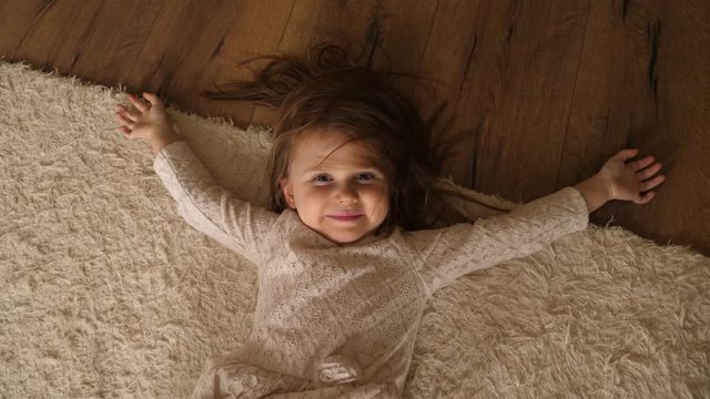 Cute little child girl play at home roll lie on a floor rug nice watch to camera