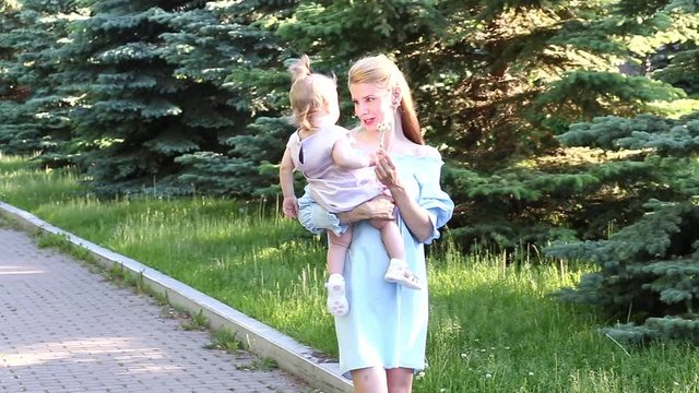 Young mother walks in the park and holds a daughter in her arms at summer day. Happy family and parenting concept