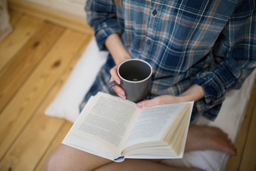 Naklejka premium Beautiful girl is sitting on the wooden floor. Young woman in men's checkered shirt is drinking coffee and reading a book on white pillow. Cozy morning with a mug in hands.