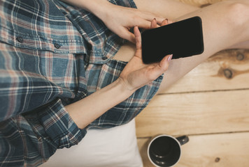 Beautiful girl is sitting on the wooden floor. Young woman in men's checkered shirt is drinking coffee and searching in social network on white pillow. Morning with black smartphone in hands.