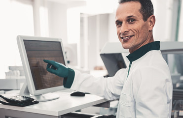 Exciting work. Portrait of pleased experienced laboratory assistant wearing green gloves and pointing at screen while searching for patients medical history