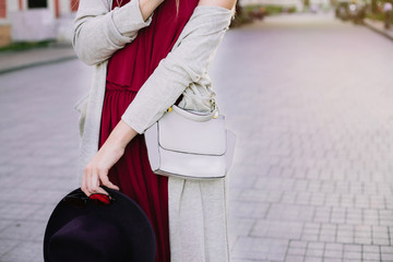 Stylish beautiful woman walking in street, wearing bordo dress, holding purse, beige jacket,sunglasses and wide hat, fashion outfit, autumn trend, accessories, hands close-up, details. Copy space