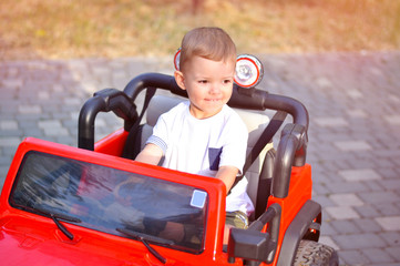 Cute 2 year old boy in a white T-shirt is riding a red jeep in the park