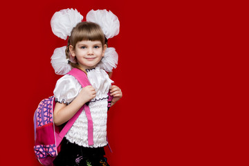 Cute smiling little girl in school uniform and white bows with backpack on red background. Back to school. Education and school concept