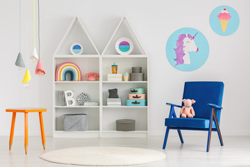 Plush toy on a blue armchair and colorful pendant lamps above an orange table with crayons legs in a cute child bedroom interior