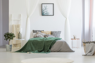 Green blanket thrown on double bed with many pillows and grey sheets standing in white hotel room interior