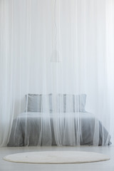 Fototapeta na wymiar Mosquito net bed with grey bedding standing in white room interior with round rug on the floor