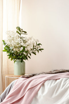Photo with bed with pink blanket in blurred foreground and bedside table with flowers in green ceramic vase standing in white bedroom interior