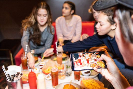 Cropped image of teenage boy sitting with friends while photographing food and drinks on table at restaurant