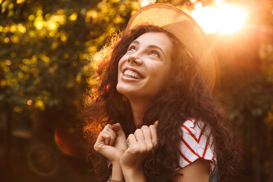 Photo Closeup Of Brunette Smiling Woman 18-20 Wearing Straw Hat Looking Upward, While Walking Through Park In Sunny Day With Trees Background