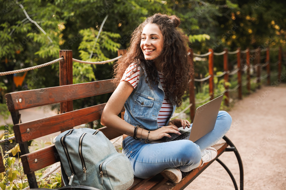 Poster photo of pretty young woman 20s with curly hair sitting on bench in green park on sunny day, and stu