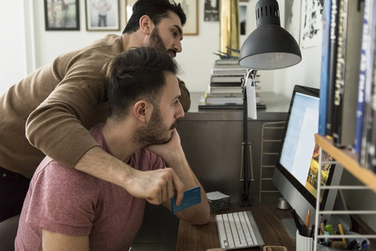 Gay Couple Shopping Online Through Credit Card On Computer At Home