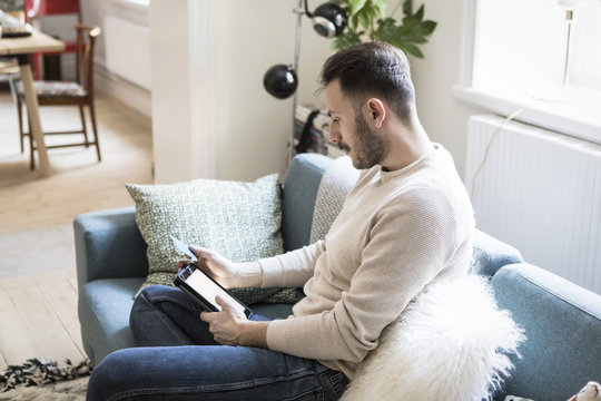 Young man doing online shopping on digital tablet through credit card while sitting on sofa at home