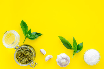 Composition with green pesto sauce in glass jar, basil leaves and garlic on yellow background top view copy space