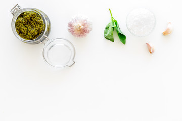 Green pesto sauce in glass jar near basil leaves and garlic on white background top view copy space