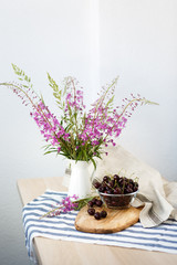 There is a plate with a cherry and a bouquet of field pink flowers in a white vase