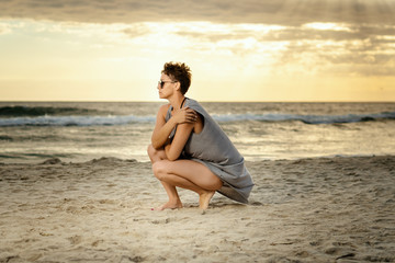 Fototapeta na wymiar beautiful woman in a gray dress crouches on the sand at the beach at sunset