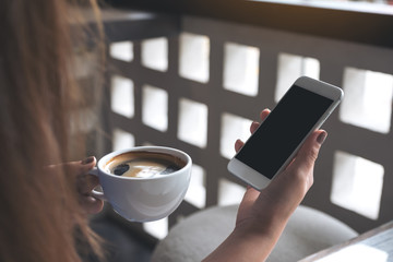Mockup image of woman's hands holding white mobile phone with blank black screen while drinking coffee in cafe