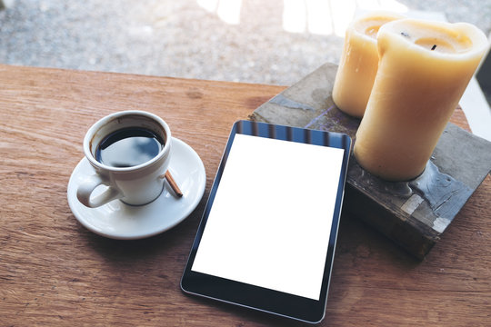 Top view mock up image of black tablet with white blank desktop screen and coffee cup , book , candles on vintage wooden table in cafe