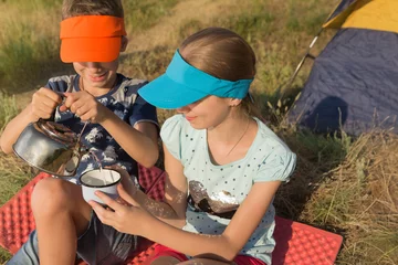 Tuinposter two children, a boy and a girl are sitting on a red rug near the tent, resting and drinking water, the boy pours water into a cup from the teapot, smiling, a summer trip, an exciting journey © aneduard