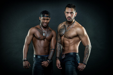 Fototapeta na wymiar Machos with muscular tattooed torsos look attractive, dark background. Masculinity concept. Guys sportsmen with sexy muscular torsos. Athletes on confident faces with nude muscular chests