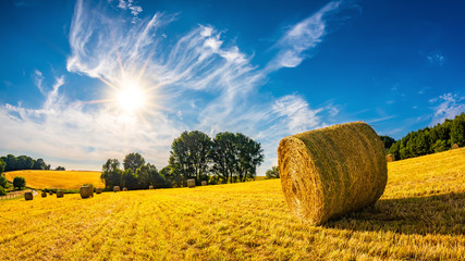 Landschaft im Sommer mit Heuballen auf einem Feld und blauem Himmel mit strahlender Sonne im Hintergrund
