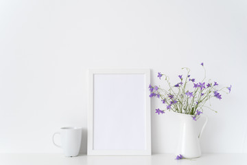 White portrait frame mockup with wild flowers in vase near white wall. Empty frame mock up for presentation design. Template framing for modern art.