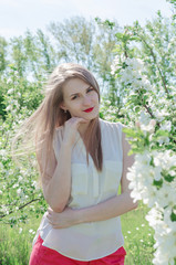 Portrait of a young beautiful woman in white top standing among blooming apple trees on a sunny day.