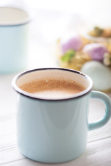 Latte in a blue enamel cup with colorful eggs and flowers in the background. Close up. Spring.