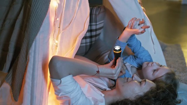 Tilt down shot of caring mother and cute little daughter lying on pillow in teepee play tent and playing with flashlight