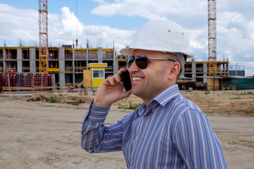 The developer in white helmet on the construction site speaks on a mobile phone and smiles. The...