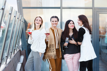 Four young charming women classmates celebrating ten years after graduation with glasses of wine on the open roof of a beautiful and expensive modern restaurant