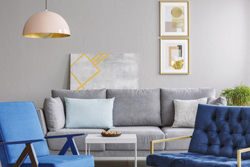 Real photo of a pink lamp hanging above blue armchairs, gray sofa and white table in cozy living...