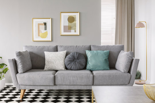Close-up of a gray sofa with white and mint cushions on a checkered floor in classy living room interior with posters on gray wall. Real photo