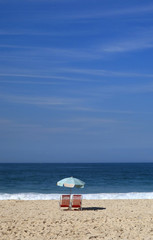 Pair of red beach chair and a blue beach parasol  on the sandy beach facing blue ocean and sky 