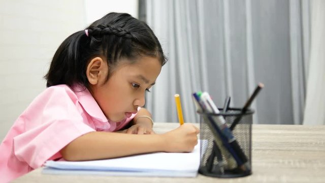 the little girl is doing homework happily. Children use a yellow pencil is writing a notebook on the wooden table.