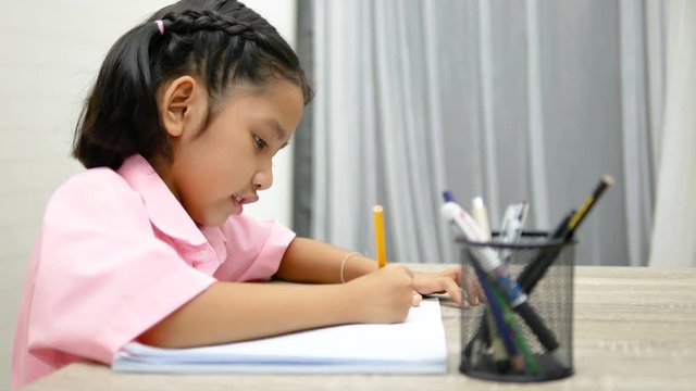 the little girl is doing homework happily. Children use a yellow pencil is writing a notebook on the wooden table.